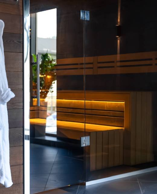 A white robe hangs by the glass sauna door, warm backlight on timber and marble sets a clean eco mood. A slow living digital detox visual for modern hotel wellness stories.